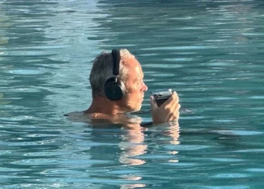 Gray-haired man neck-deep in turquoise pool water wearing over-ear headphones and holding a smartphone